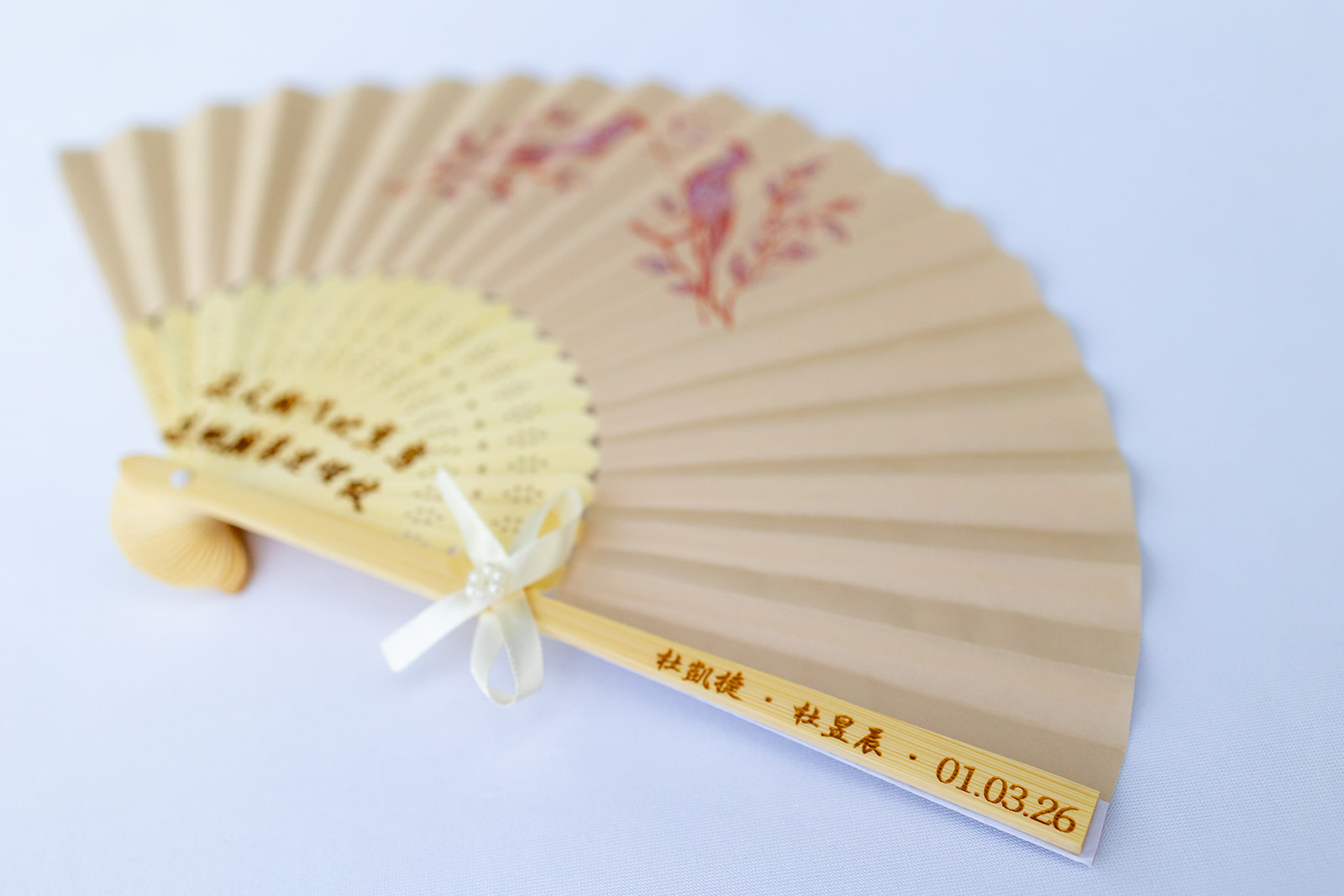 A beige hand fan with red floral and bird designs, featuring a ribbon bow and text including a date, against a plain white background.