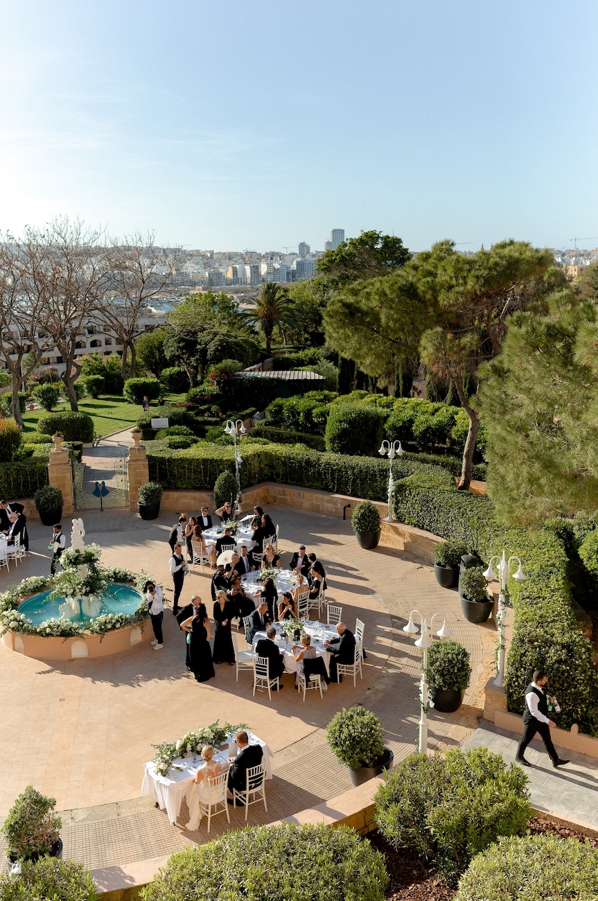 People are seated at round tables near a small fountain in an outdoor garden setting, surrounded by greenery and trees, with a city skyline visible in the background.