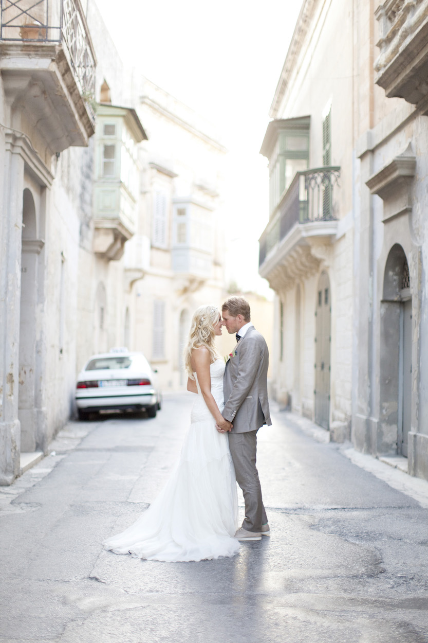 A bride and groom stand facing each other in the middle of a quiet, sunlit street lined with old buildings.
