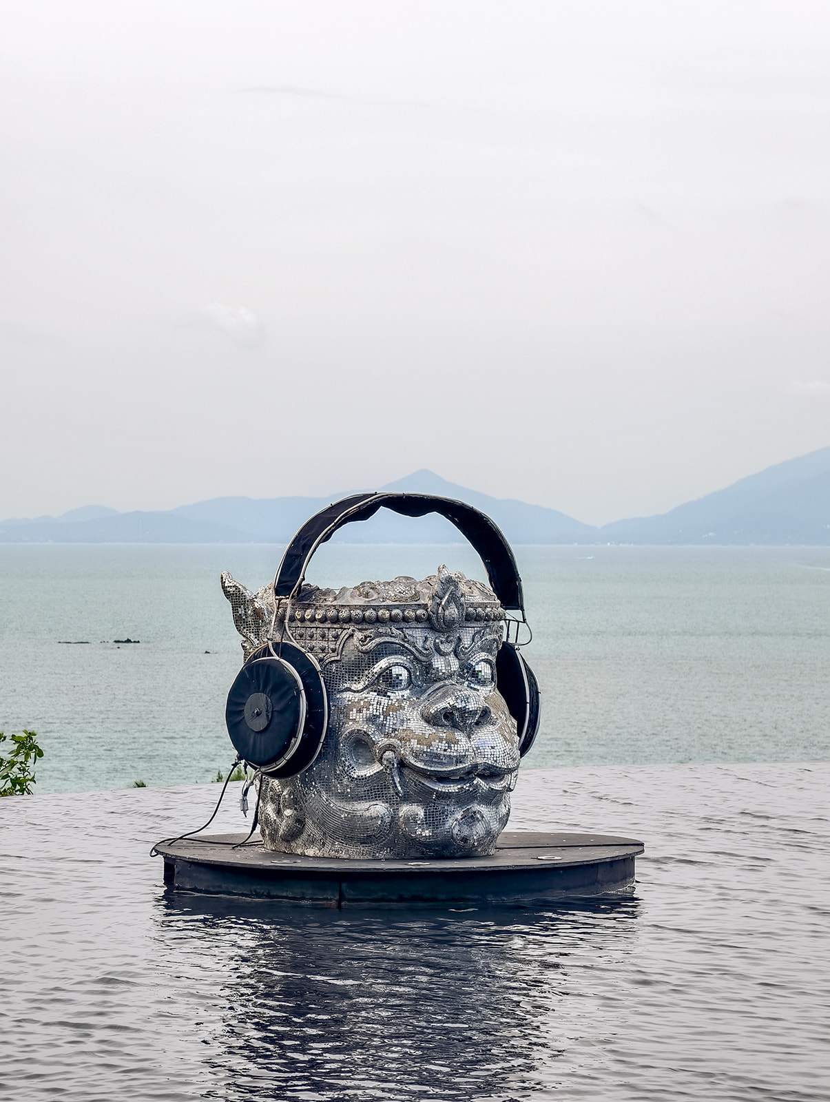 A large stone sculpture of a face wearing oversized headphones sits on a circular platform by the water, with a calm sea and mountains in the background.