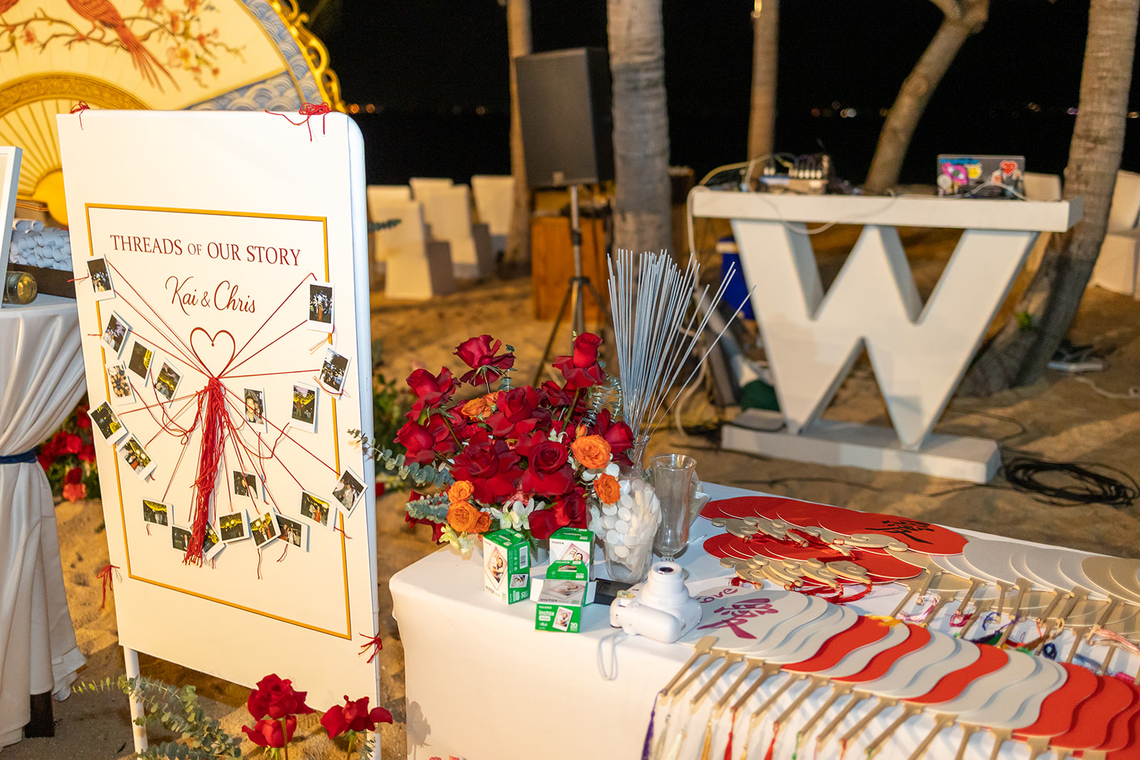 Outdoor event setup at night with a decorated table, a display board with photos, flowers, and a large white "W" structure in the background.