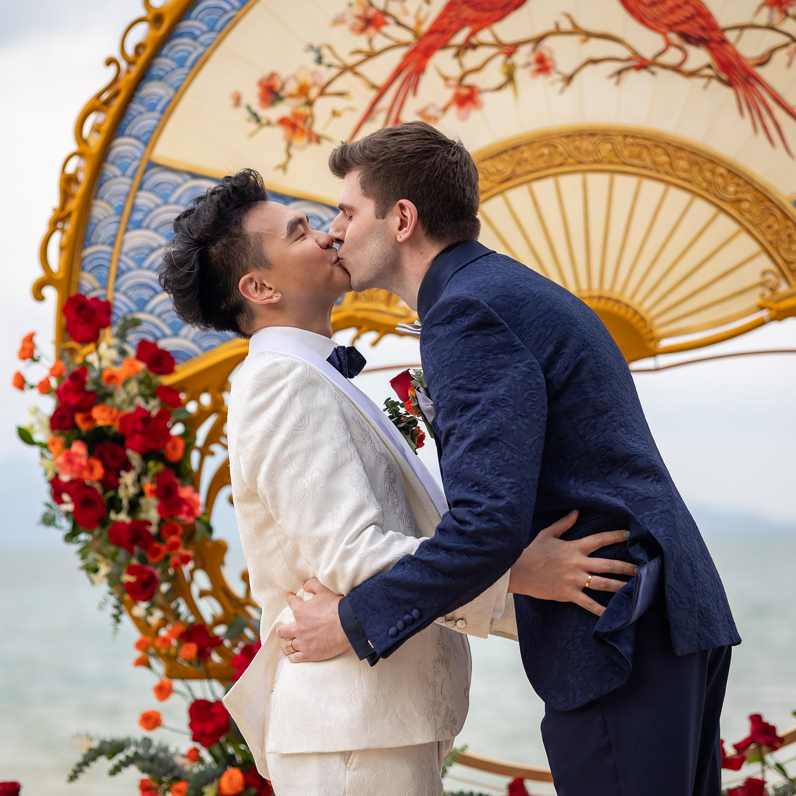 Two grooms kiss in front of a floral arch and decorative fan with red birds during an outdoor wedding ceremony by the water.