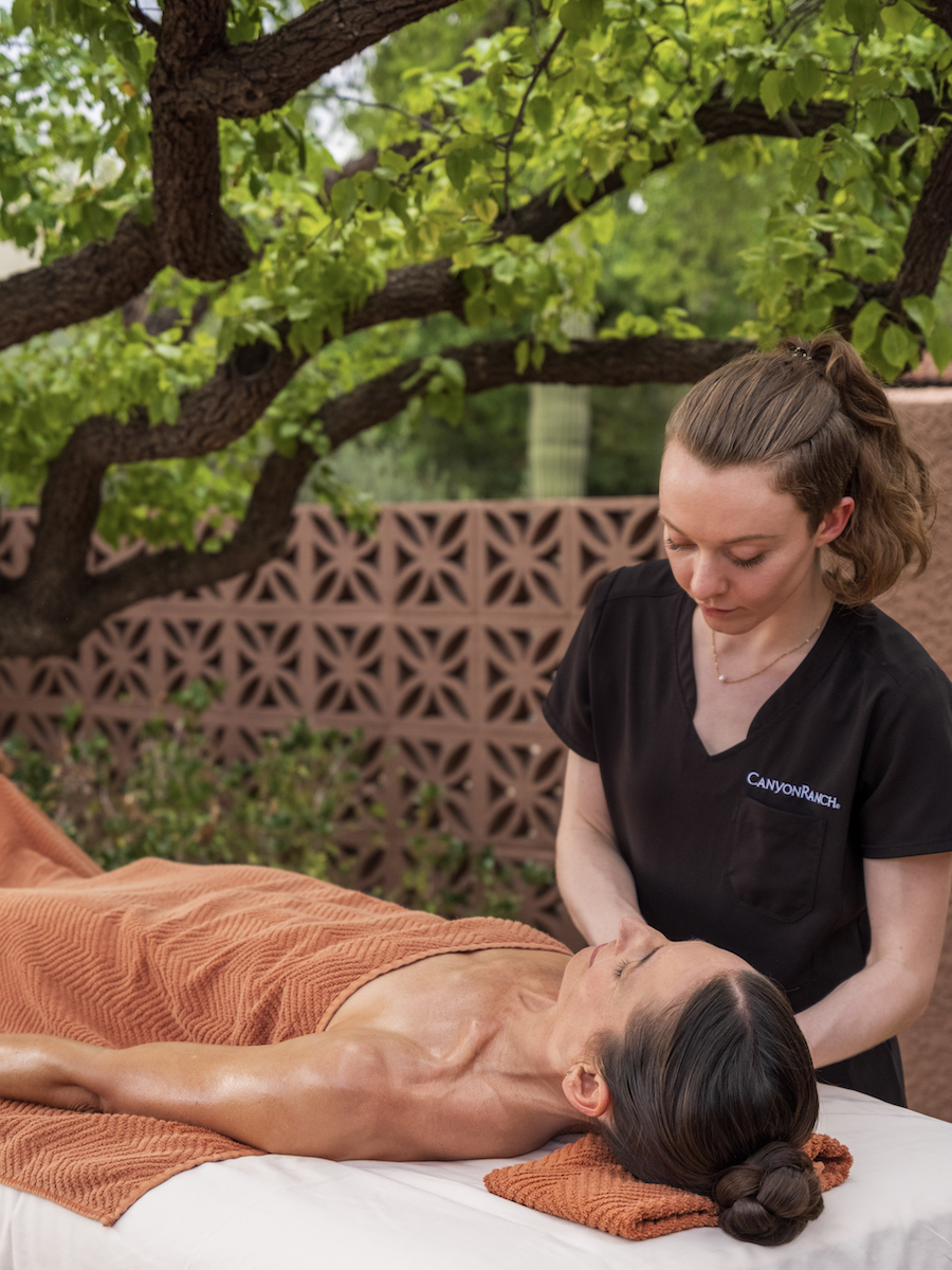 A massage therapist gives a massage to a person lying on a table outdoors, with trees and a brick wall in the background.