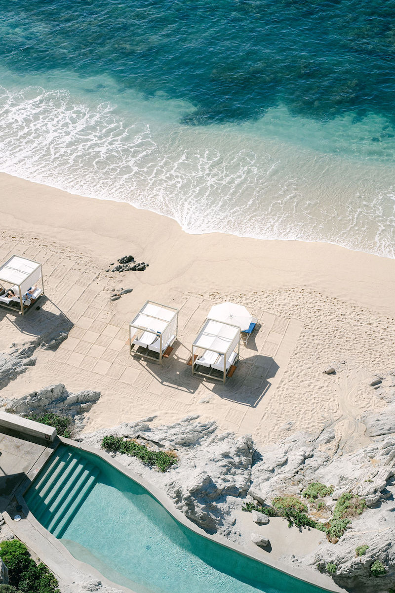 Aerial view of a sandy beach with three white cabanas, clear blue ocean waves, and a pool with stone steps in the foreground.
