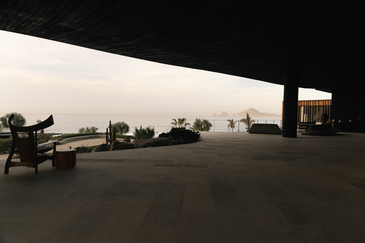 Spacious covered patio with minimalist furniture overlooks palm trees, garden, and a hazy ocean view under a wide overhang.
