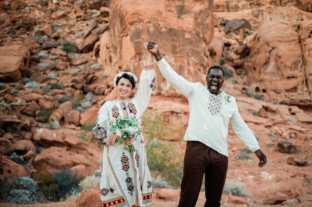 A smiling couple in embroidered clothing stands in a rocky desert landscape, holding hands up in celebration. The woman holds a bouquet and wears a flower crown.