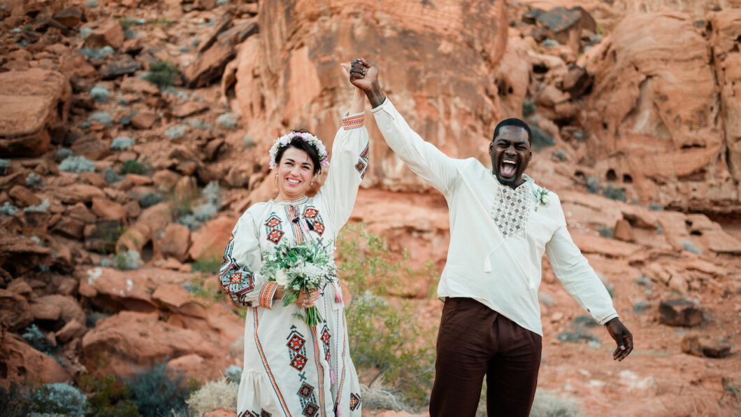 A smiling couple in embroidered clothing stands in a rocky desert landscape, holding hands up in celebration. The woman holds a bouquet and wears a flower crown.