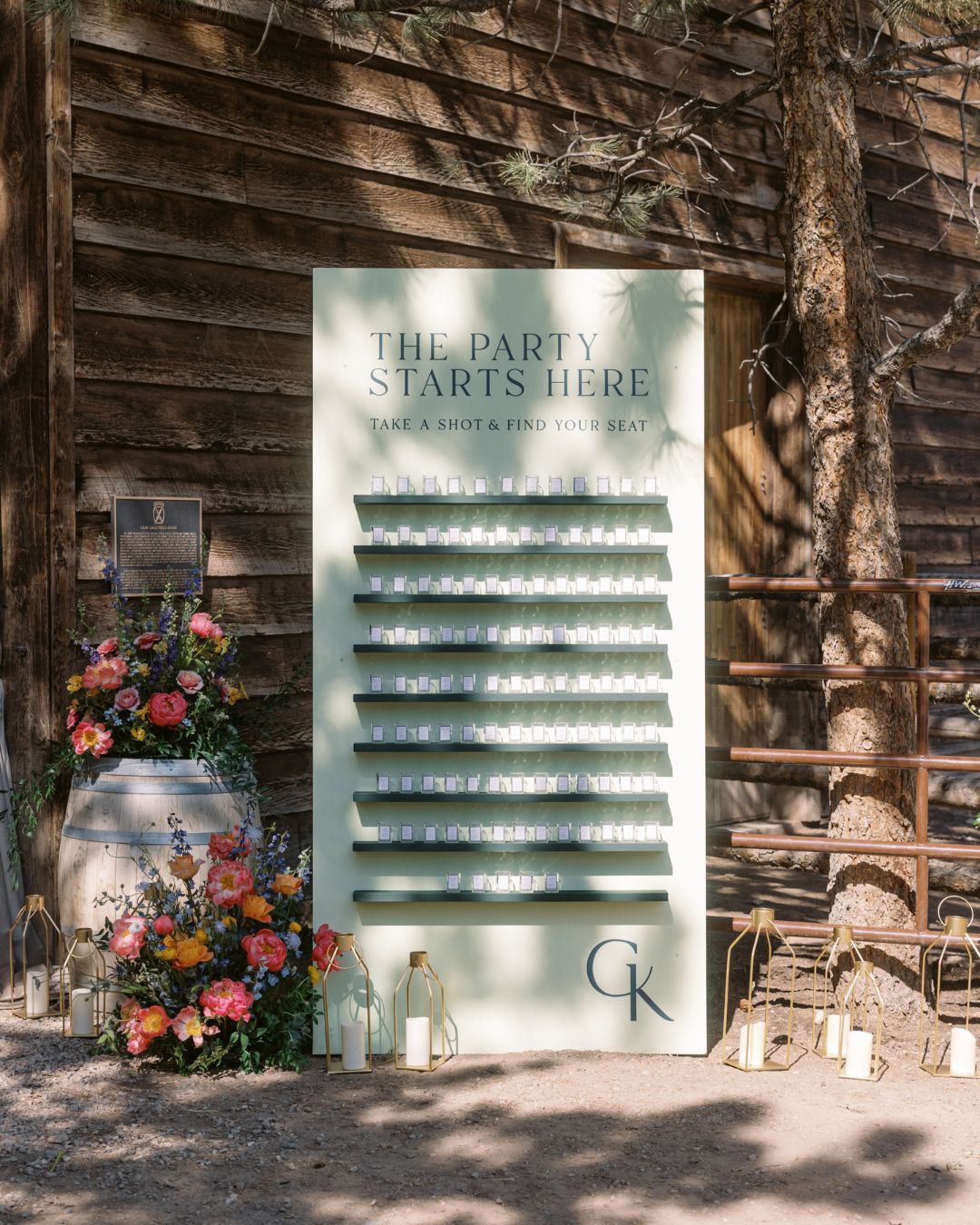 A display board labeled "The Party Starts Here" holds rows of clear glasses, with flowers and bottles arranged at the base, set outdoors beside a rustic wooden wall.
