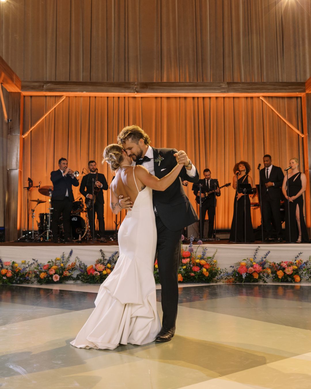 A bride and groom share a first dance on a ballroom floor, with a live band and floral arrangements in the background.