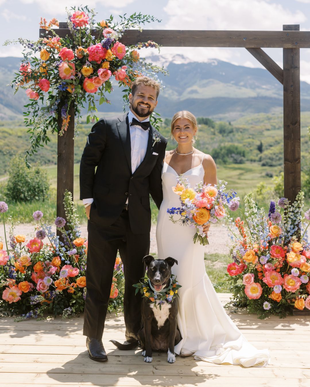 A bride and groom stand outdoors with a dog in front of a floral arch, surrounded by colorful flowers and mountains in the background.
