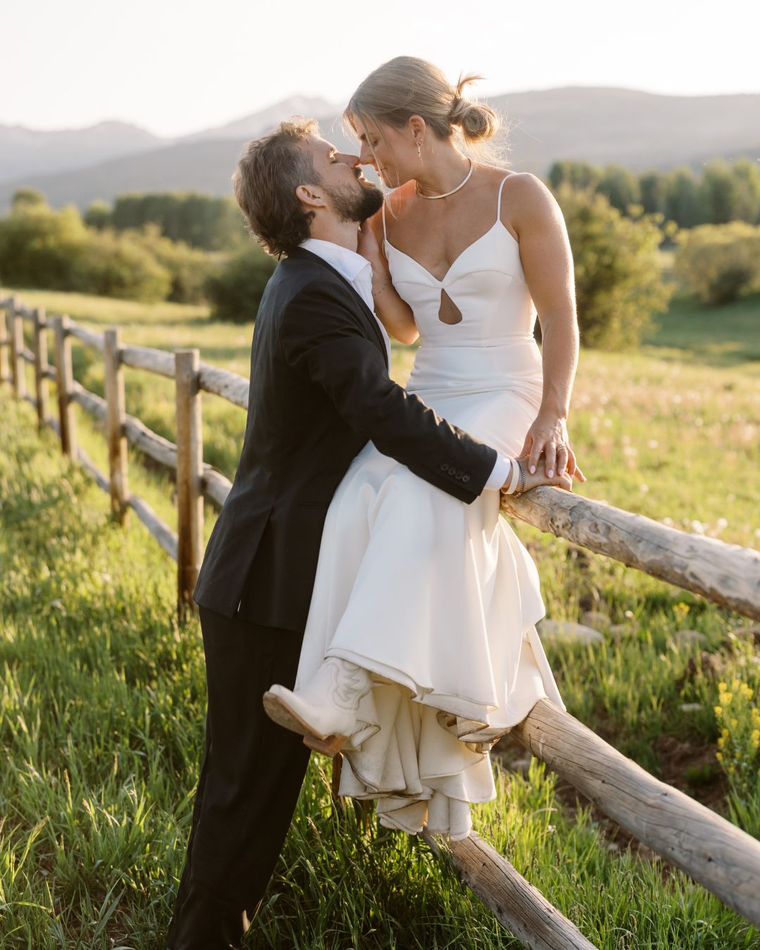 A bride in a white dress sits on a wooden fence while a groom in a black suit stands close, outdoors in a grassy field with trees and mountains in the background.