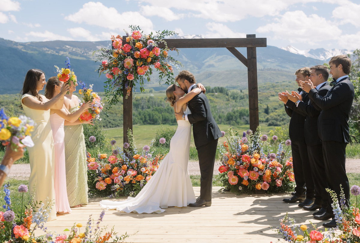 A bride and groom embrace and kiss on an outdoor wooden platform decorated with colorful flowers, surrounded by applauding wedding party members, with mountains and greenery in the background.