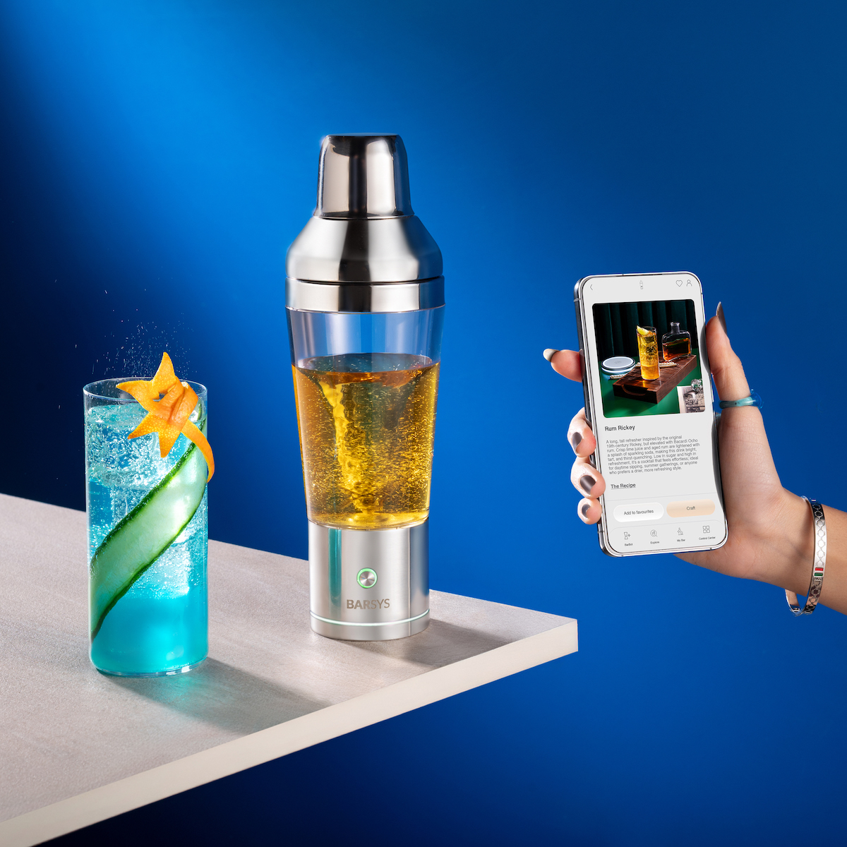 A hand holds a smartphone displaying a cocktail recipe next to an electric cocktail shaker and a blue cocktail with a starfruit and cucumber garnish on a white table against a blue background.