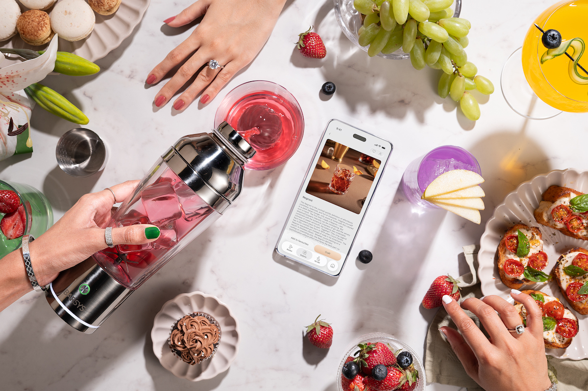 A table with snacks, fruit, drinks, and a smartphone displaying a cocktail recipe; two hands are preparing and enjoying food and beverages.