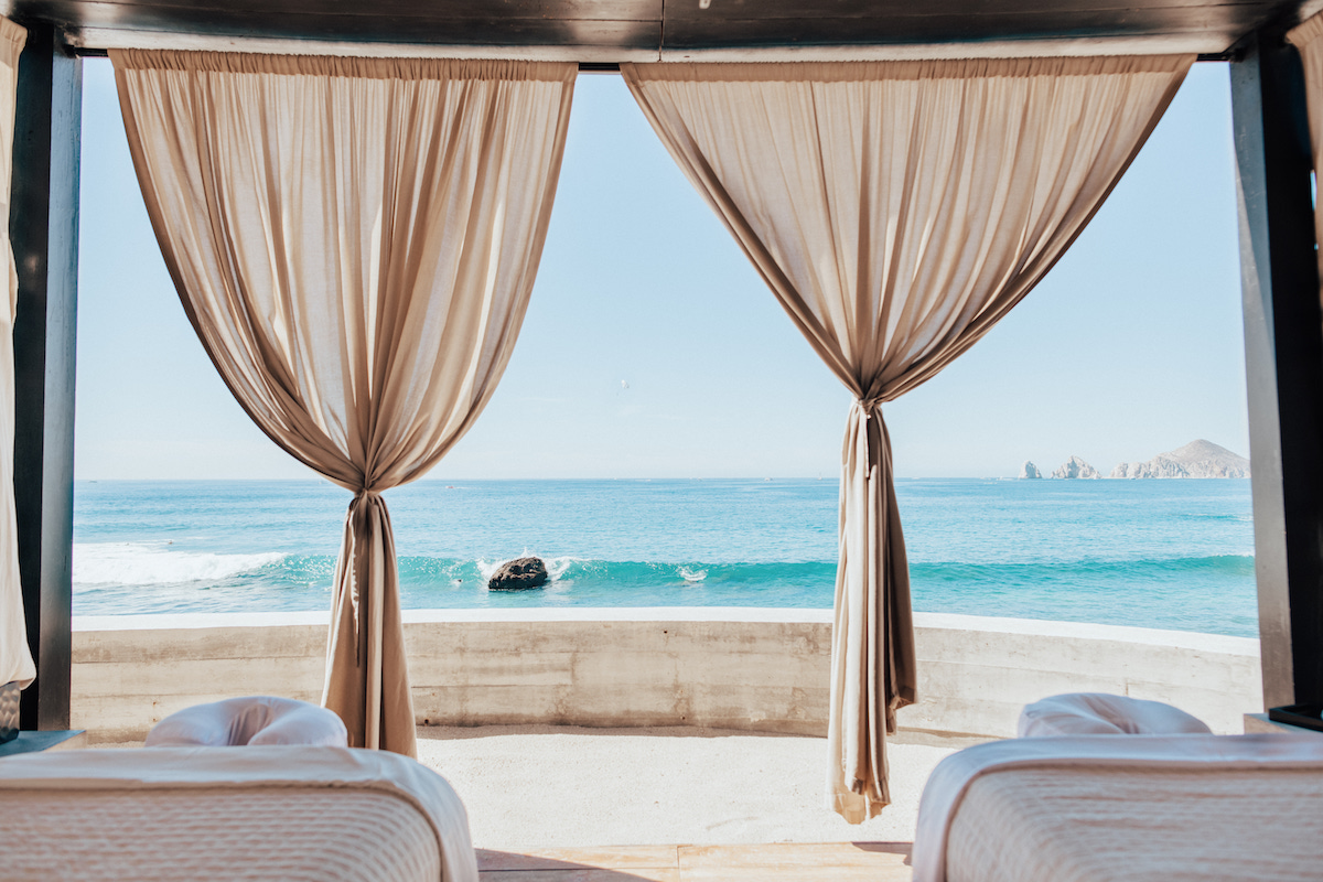 Two massage tables under a cabana with open beige curtains overlook a calm ocean and clear blue sky.