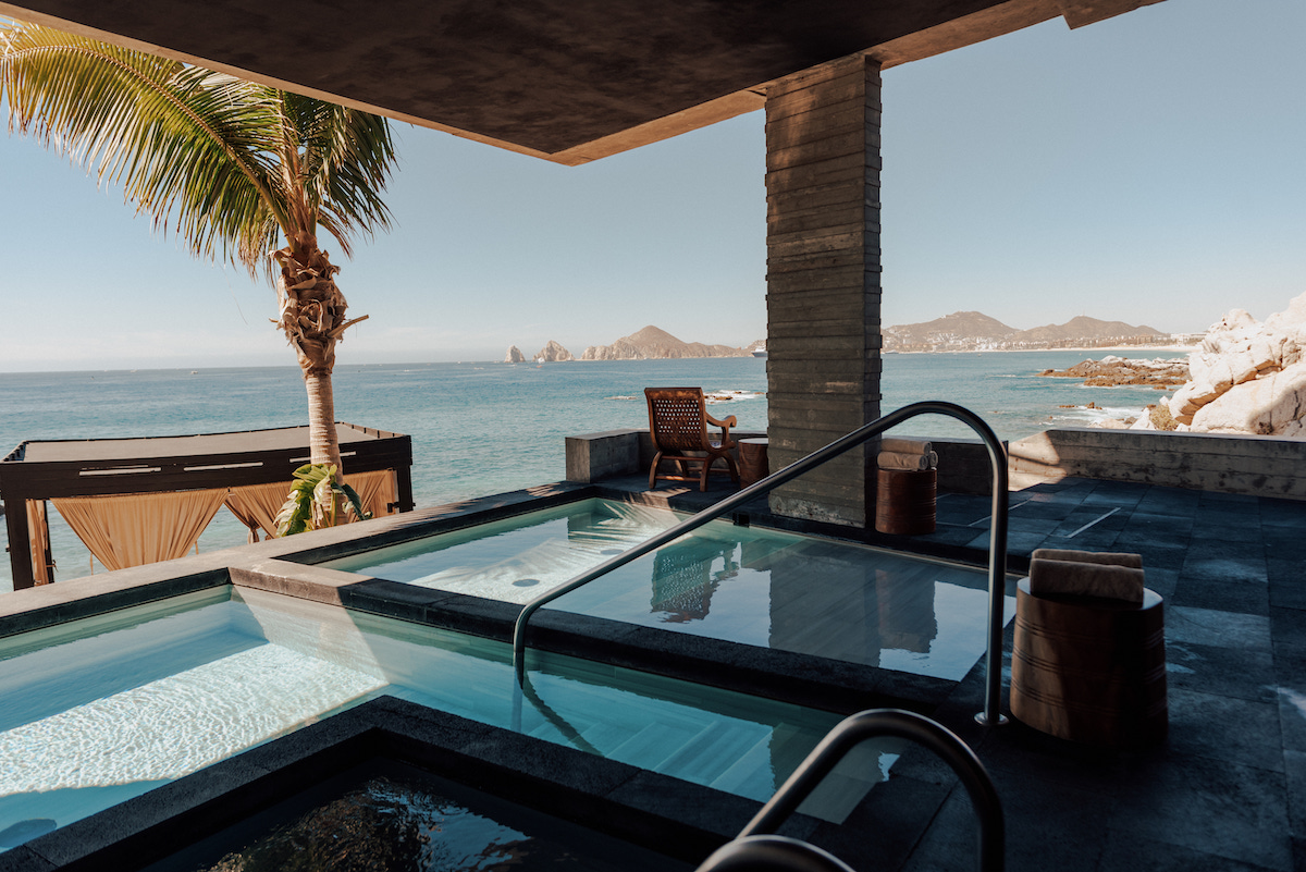 Outdoor hot tubs overlooking the ocean, with a palm tree, lounge chairs, and distant mountains under a clear sky.