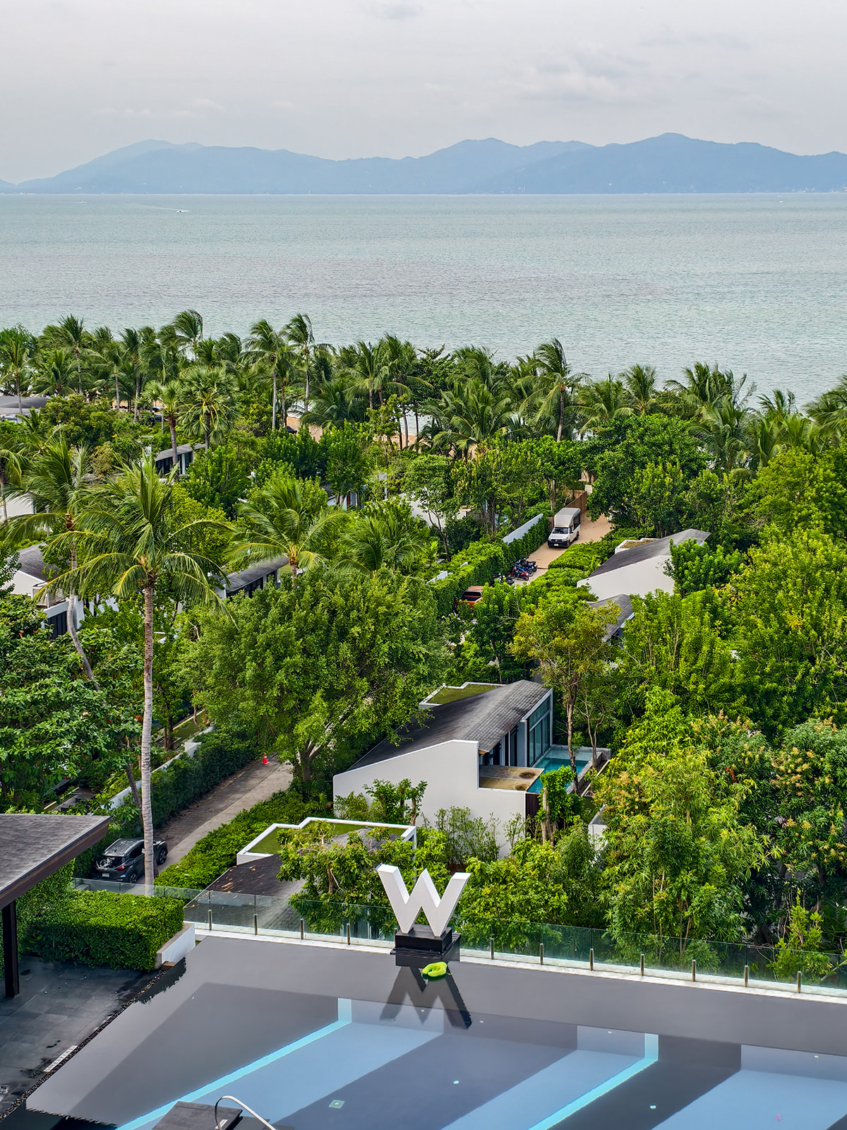 A rooftop with a large "W" logo overlooks lush greenery, a coastline, and distant mountains under a cloudy sky.