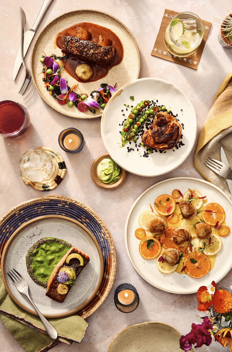 An overhead view of a table set with four artfully plated gourmet dishes, two drinks, candles, silverware, and a small vase with flowers.