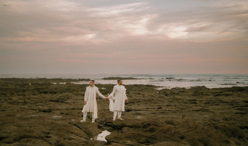Two people dressed in white stand holding hands on a rocky shoreline at sunset, with the ocean and a cloudy sky in the background.