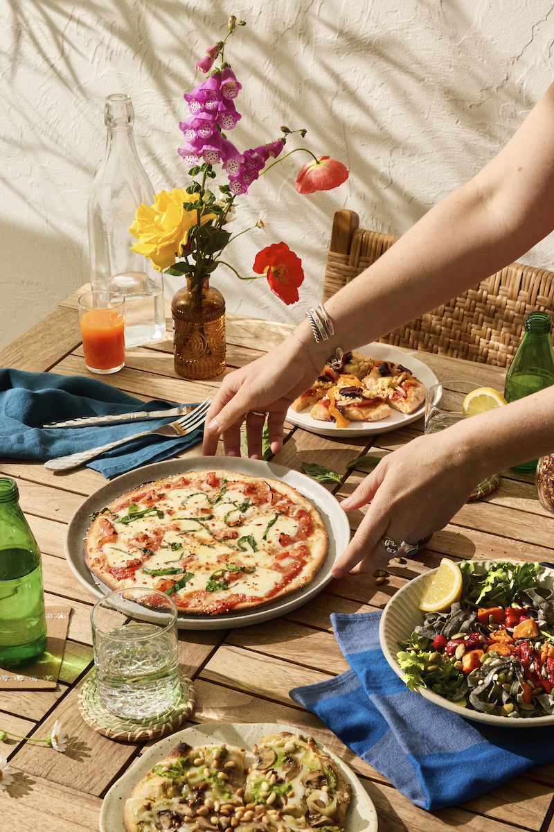 A person sets a pizza on a wooden table with other dishes, drinks, and a vase of colorful flowers in natural sunlight.
