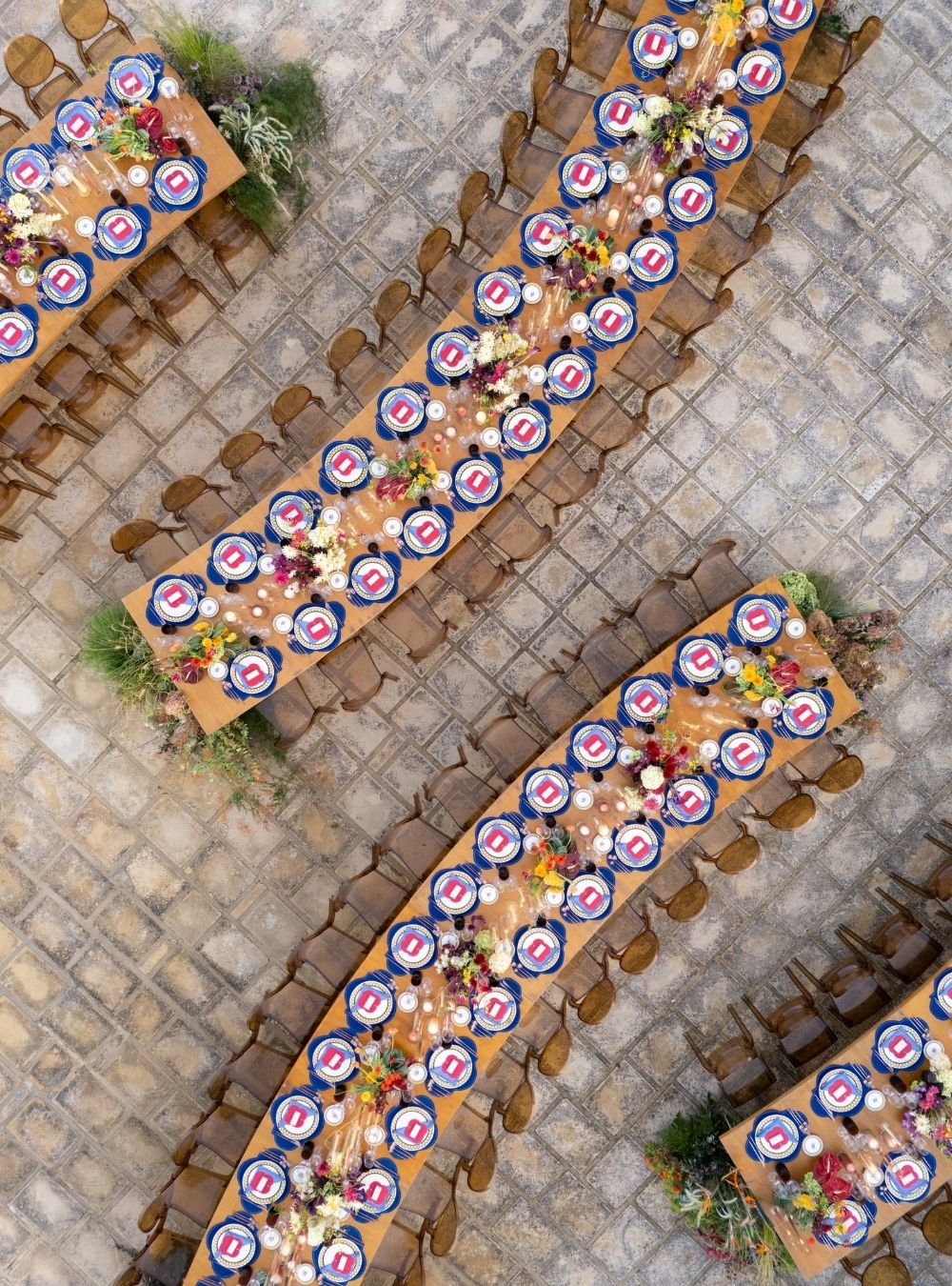 Aerial view of long banquet tables set with plates, cutlery, and floral centerpieces, arranged in curved rows on a cobblestone surface.