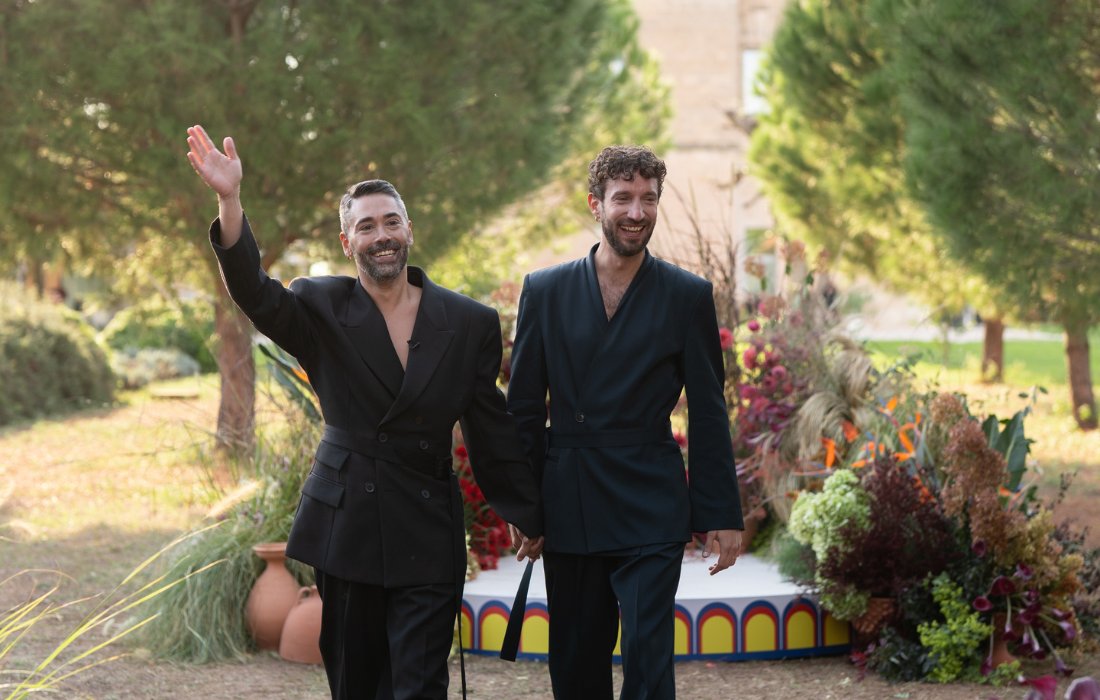 Two men in black suits hold hands and smile while walking outdoors, with one waving. Decorative plants and a small stage are in the background.