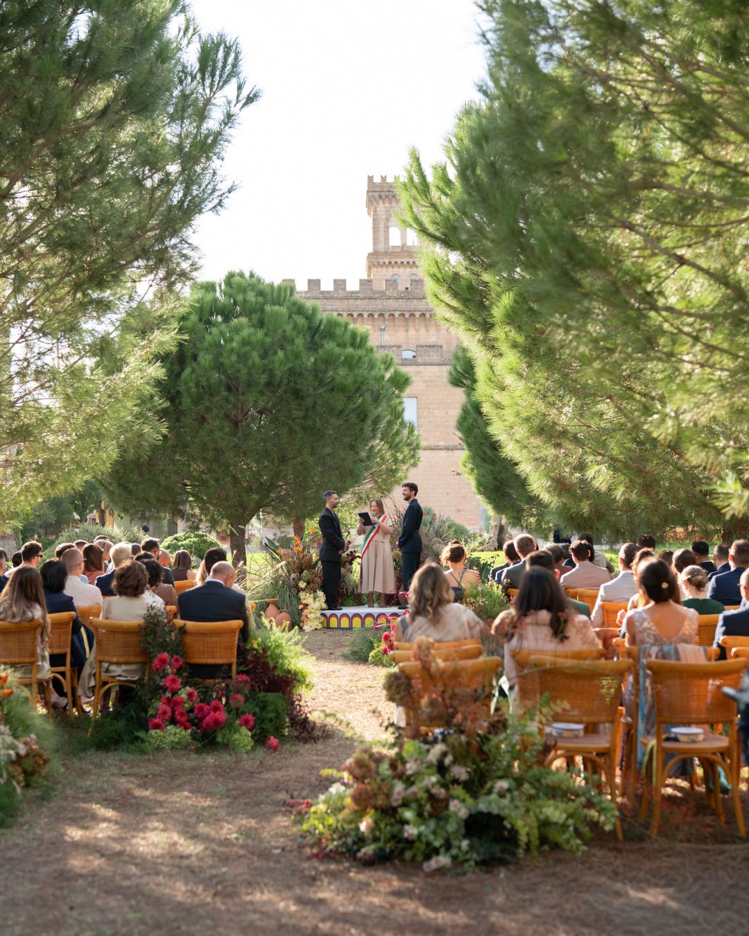 Outdoor wedding ceremony with guests seated on wooden chairs, a couple standing at the altar, and trees surrounding the area. A large building is visible in the background.