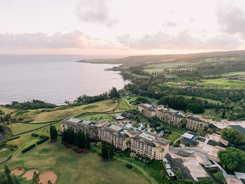 Aerial view of a large coastal resort surrounded by golf courses and greenery, with the ocean and a hilly landscape in the background at sunset.