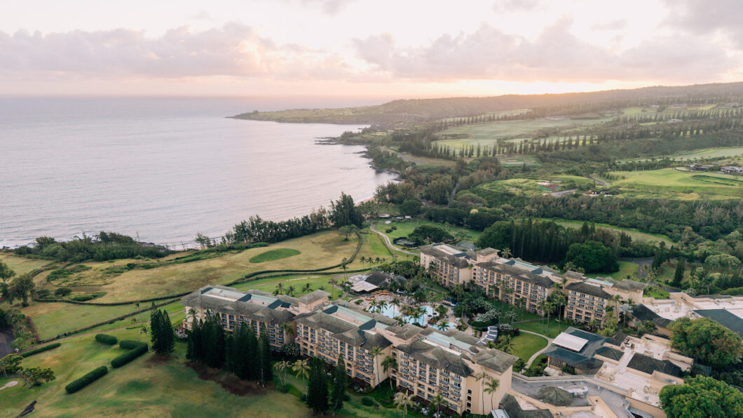 Aerial view of a large coastal resort surrounded by golf courses and greenery, with the ocean and a hilly landscape in the background at sunset.