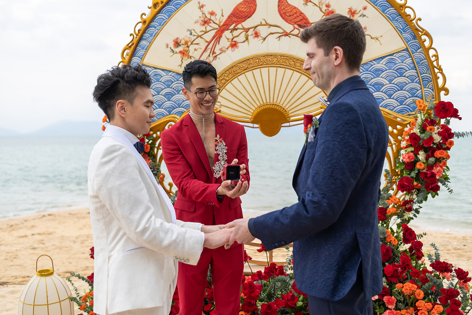 Two men stand facing each other holding hands on a beach while an officiant holds a ring box. A decorative fan and red flowers are displayed in the background.