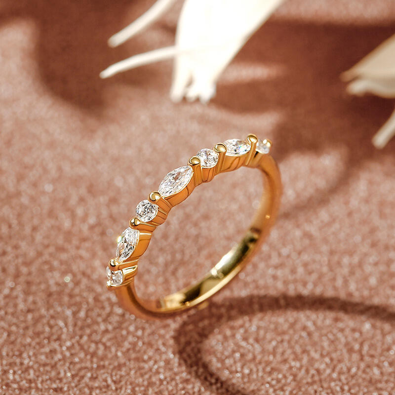 Gold ring with alternating round and marquise-cut clear stones, displayed on a textured brown surface with decorative white accents in the background.
