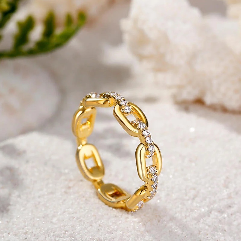 A gold chain-link ring with embedded small diamonds is displayed on a sandy surface with white coral and green foliage in the background.