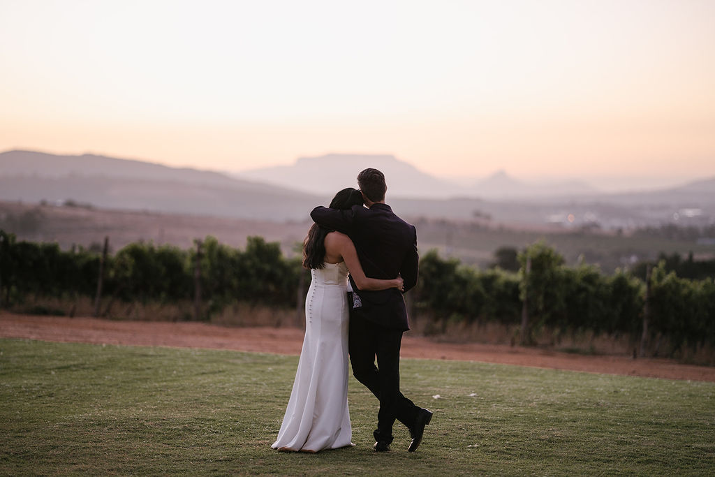 A couple dressed in formal attire stands arm in arm on a lawn, facing a vineyard and distant mountains at sunset.