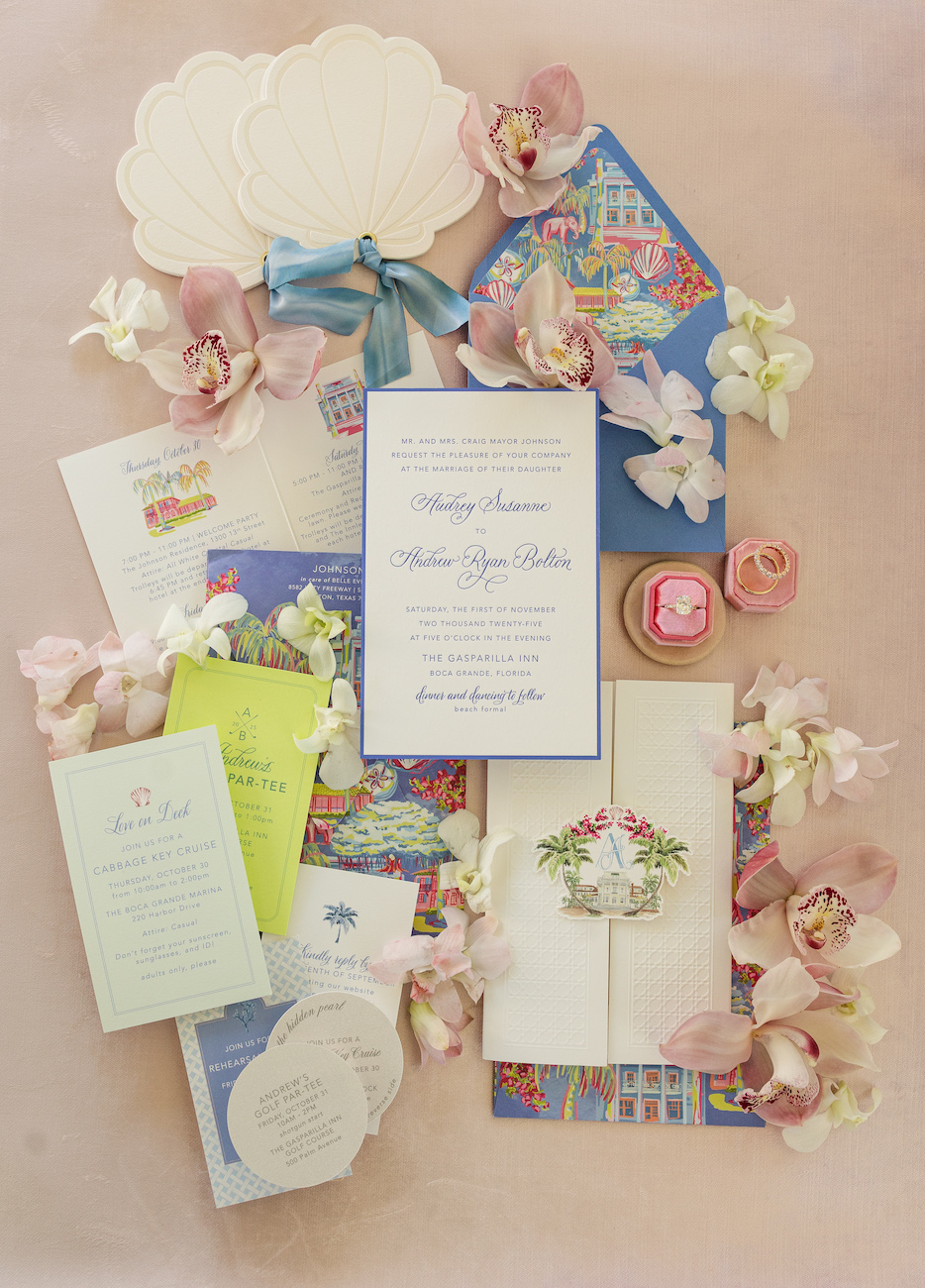 A flat lay of colorful wedding invitations, envelopes, a ring box with rings, and scattered orchid flowers arranged on a light pink surface.