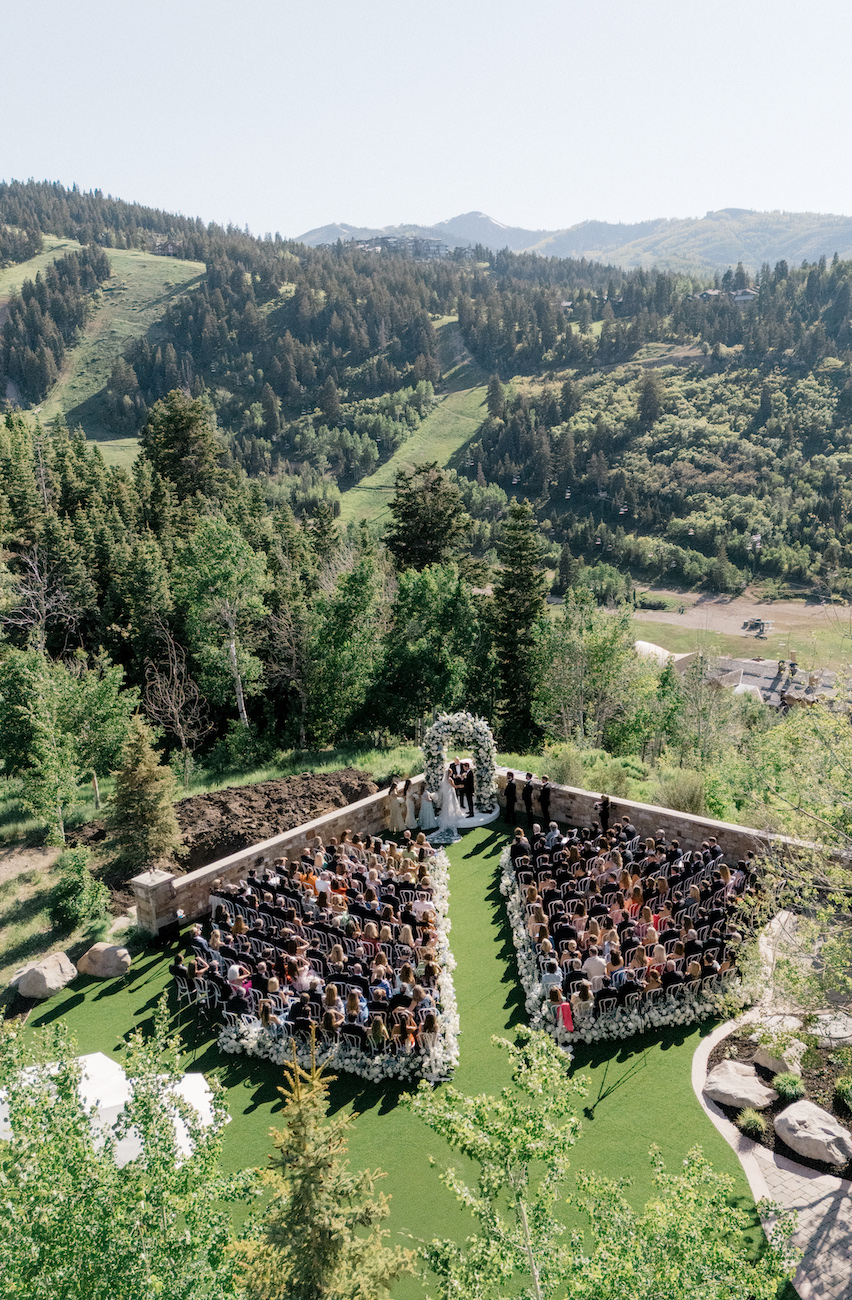 An outdoor wedding ceremony is taking place on a lawn surrounded by trees and mountains, with guests seated facing a floral arch where the couple stands.
