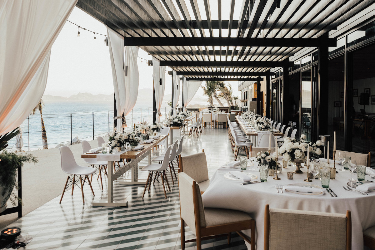 Covered outdoor dining area set for an event with white tablecloths, floral centerpieces, and ocean view in the background.