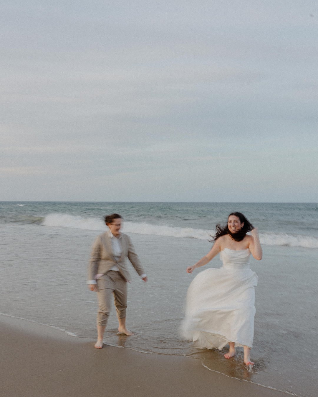 Two people in formal attire, one in a dress and one in a suit, walk and smile barefoot along the shore of a beach with gentle waves in the background.