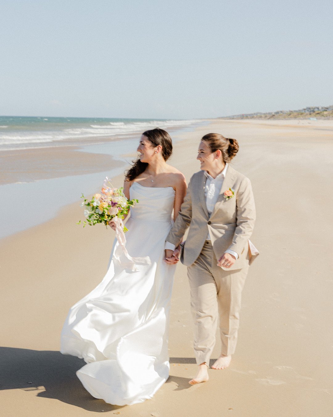 Two women, one in a wedding dress and one in a light suit, walk barefoot holding hands on a sandy beach, with the ocean and sky in the background.