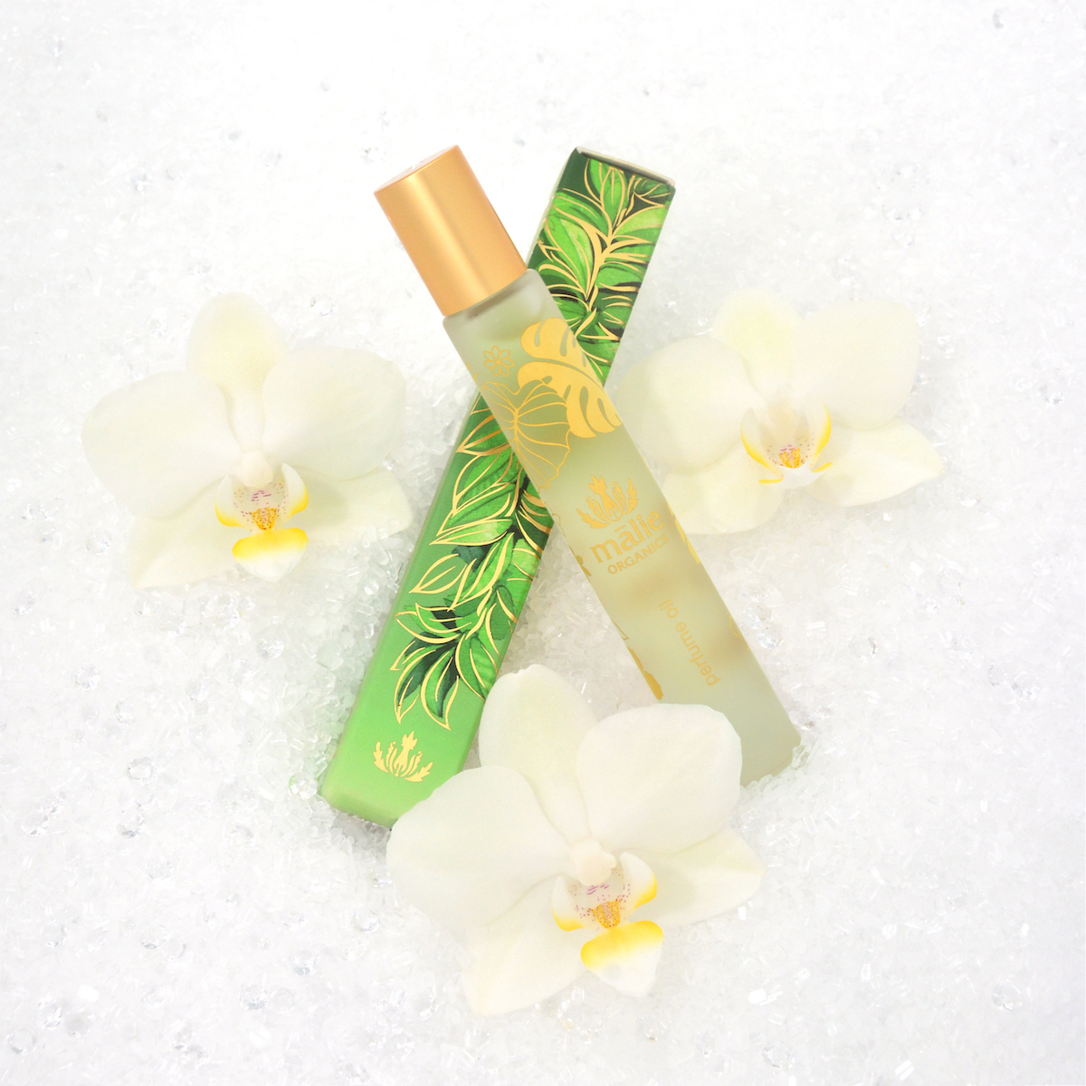 A perfume bottle with a gold cap is placed next to a green leafy box, surrounded by three white orchid flowers on a white textured surface.