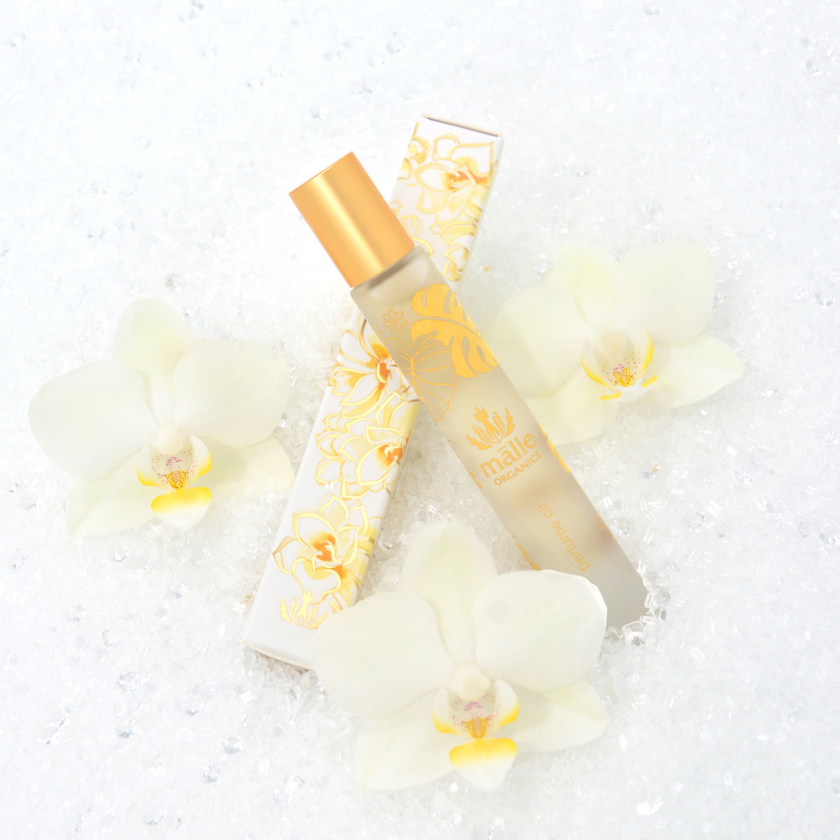A frosted glass perfume bottle with a gold cap and floral design lies next to its matching box, surrounded by three white orchids on a white textured surface.