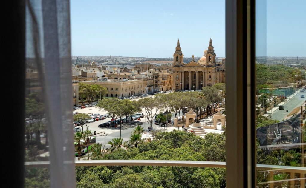 View through a window overlooking a city square with trees, parked cars, and a large historic church with two bell towers, under a clear sky.