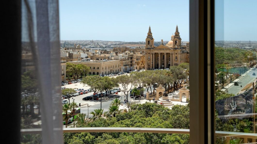 View through a window overlooking a city square with trees, parked cars, and a large historic church with two bell towers, under a clear sky.