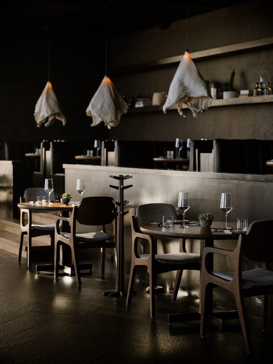 A modern restaurant interior with wooden tables and chairs, set with glassware, and pendant lights with fur coverings hanging from the ceiling.