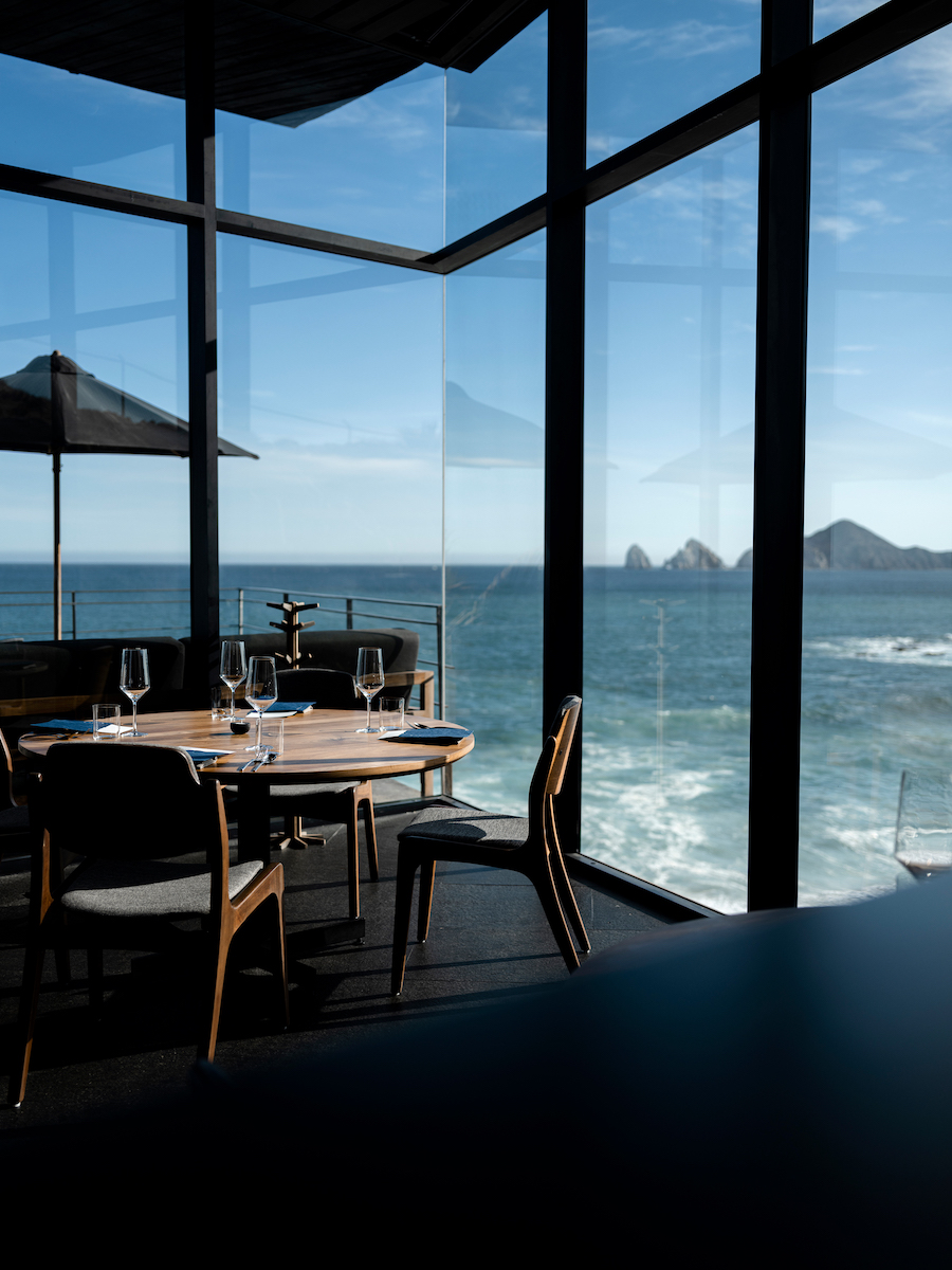 A dining table set for four is positioned by large windows overlooking the ocean and distant rocky formations under a clear sky.