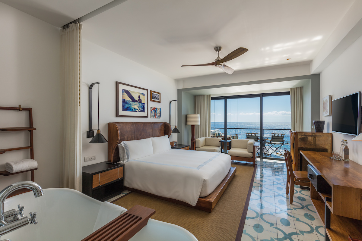 Modern hotel room with a king-size bed, desk, bathtub, seating area, and large windows overlooking a balcony with an ocean view.