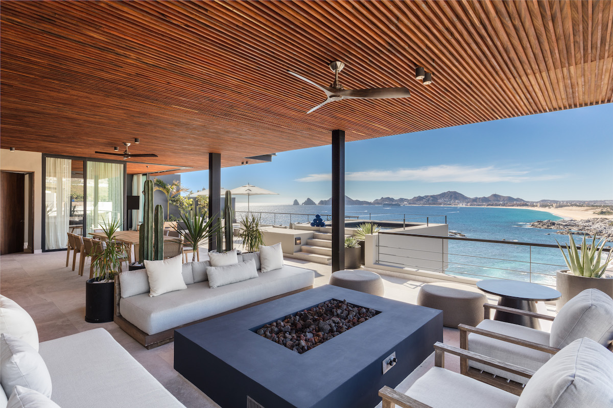 Modern outdoor lounge with white sofas and a fire pit, covered by a wooden ceiling, overlooking a coastal view with blue ocean and distant mountains.