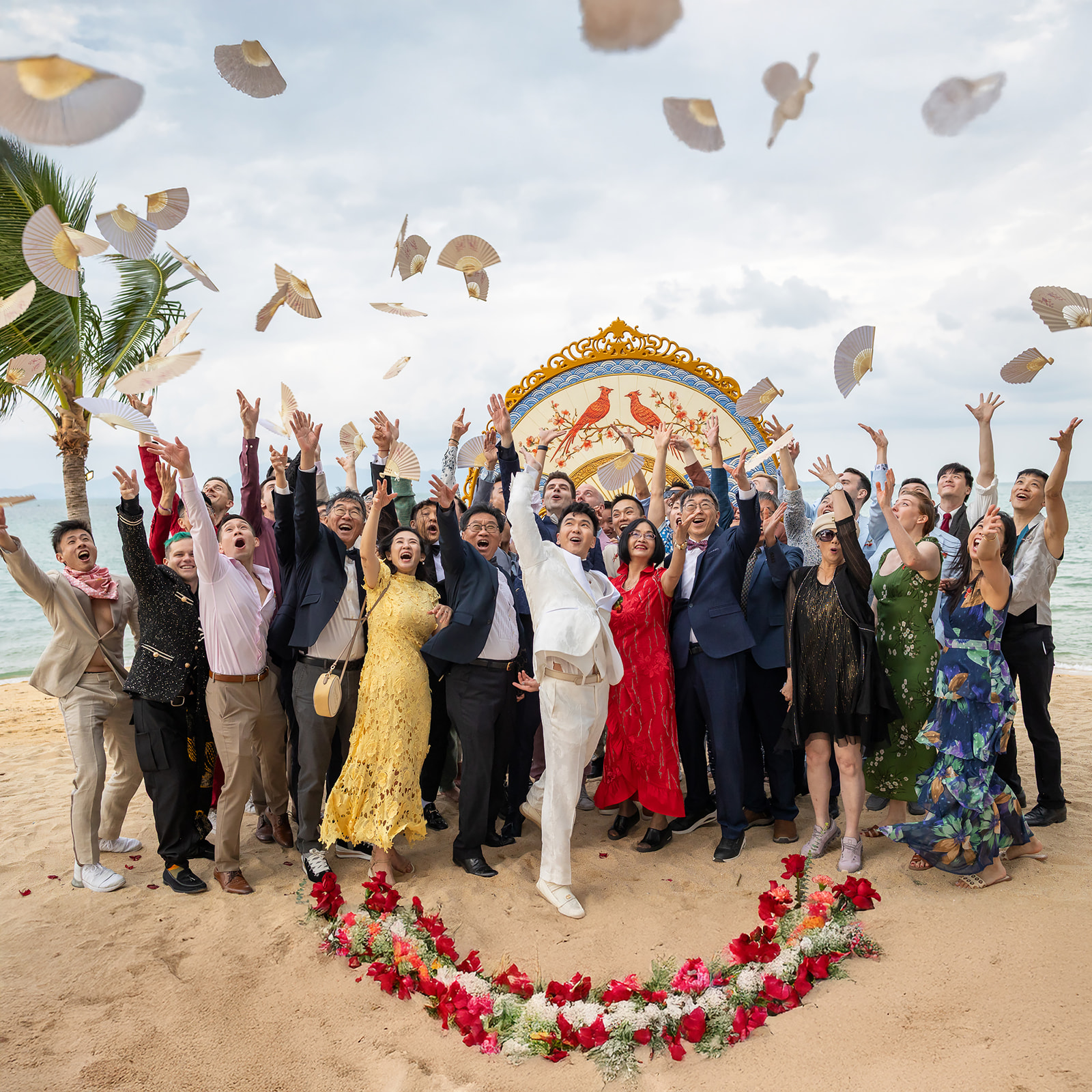 A group of people in formal attire stand on a beach, smiling and tossing paper fans into the air, with a floral heart shape on the sand in front of them.