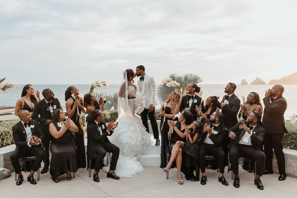 A bride and groom stand at an outdoor altar by the sea, surrounded by a wedding party dressed in black, clapping and smiling.