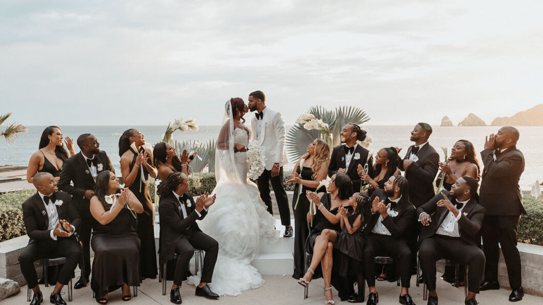 A bride and groom stand at an outdoor altar by the sea, surrounded by a wedding party dressed in black, clapping and smiling.