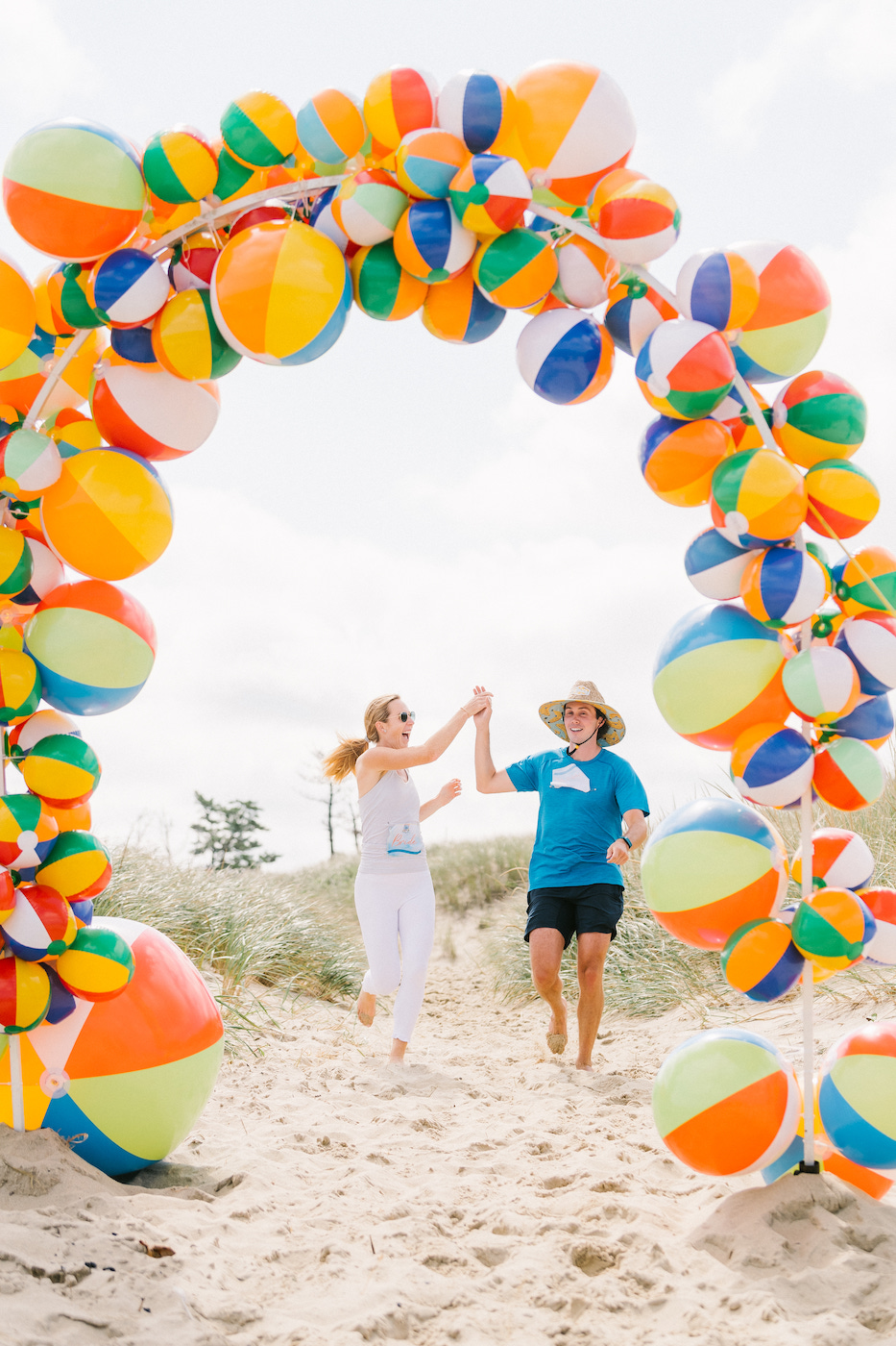 Two people hold hands and run under an arch made of colorful beach balls on a sandy path, with grass and sky in the background.